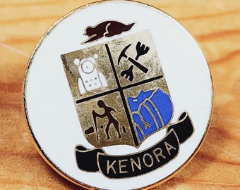 May include: A white enamel pin with a gold border featuring a crest with four sections. The crest includes a compass, a pickaxe, a hiker, and a mountain. The text "KENORA" is printed in black below the crest.