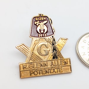 以下が含まれることがあります： 茶色のトップ部分とシンボルの上に「GIZEH」の文字が入った金色のフリーメーソンピン。ピンには「R. GLENN ALLEN POTENTATE」と年号「2013」の文字が入っています。カナダの25セント硬貨がピンの隣にあります。