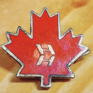 Puede incluir: Un pin de hoja de arce roja con un contorno plateado y una "H" estilizada en el centro. El pin está sobre una superficie de madera.