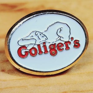 May include: A white oval pin with a gold border. The pin features a cartoon illustration of a dog sleeping with the text "Goliger's" in red letters.