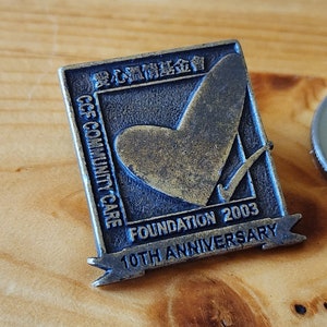 May include: A bronze-colored pin with a blue border and a heart with a checkmark inside. The text on the pin reads "CCF Community Care Foundation 2003 10th Anniversary".