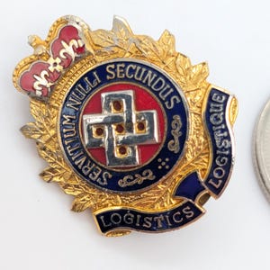 May include: A gold-toned military badge with a red, white, and blue emblem, a crown, and the words "SERVITIUM NULLA SECUNDUS LOGISTICS". The badge is next to a coin.