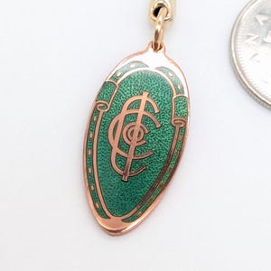 May include: A gold-toned metal keychain featuring an oval pendant with a textured green enamel inlay and a central copper-colored monogram. The keychain is positioned next to a Canadian 25-cent coin for scale, showing the relative size of the item.