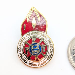 Puede incluir: Pin dorado con un diseño de llama roja sobre un emblema circular. El emblema tiene un centro azul y blanco con el texto "British Columbia Professional Firefighters Burn Fund" en un patrón circular.