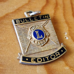 Może przedstawiać: Złota metalowa spinka z emaliowanym niebiesko-białym emblematem Lions Club i napisem "BULLETIN EDITOR" w czarnym emaliowanym kolorze.
