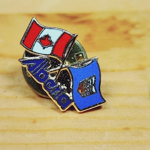 May include: A gold pin featuring a red and white Canadian flag and a blue and white Alberta flag. The pin reads "Alberta" in gold lettering.