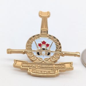Puede incluir: Un pin de solapa de metal dorado con un emblema circular central con una hoja de arce roja, rodeado por una corona de laurel. El pin incluye una barra horizontal, una forma de aleta vertical y una pancarta que dice ABBOTSFORD INTERNATIONAL AIRSHOW ESTABLISHED 1962.