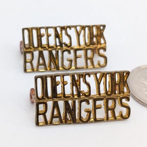 Könnte beinhalten: Zwei goldfarbene Metallanstecknadeln mit dem ausgeschnittenen Text QUEEN'S YORK RANGERS in zwei Zeilen. Eine silberfarbene Münze liegt rechts neben den Anstecknadeln als Größenvergleich und bietet einen klaren visuellen Hinweis auf die Größe.