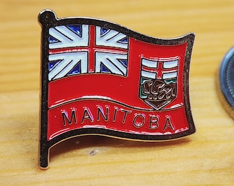 May include: A red, white, and blue enamel pin featuring the flag of Manitoba, Canada. The pin has a gold border and the word "MANITOBA" is written in gold letters.