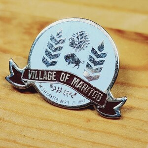 May include: Silver pin with a blue and white enamel design. The pin features a bull, wheat stalks, and the text "Village of Manitou Incorporated April 21 1891".
