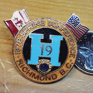 Könnte beinhalten: Eine goldene und schwarze Anstecknadel mit dem Text "1978 Spring Conference Richmond B.C." und einem blauen "H" mit der Zahl "19" darin. Die Anstecknadel ist mit einer kanadischen und einer amerikanischen Flagge verziert.