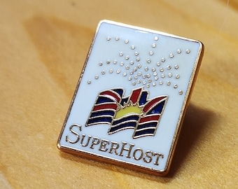 May include: A white enamel pin with a gold border. The pin features a stylized flag with a sun and the text "SUPERHOST".