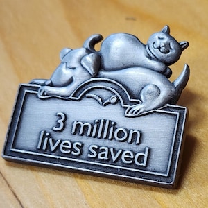 May include: Silver pin with a cat and dog cuddling on a sign that reads "3 million lives saved".
