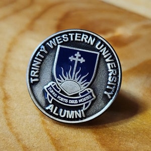 May include: Silver Trinity Western University alumni pin with a blue and silver shield featuring a cross and sun. The pin reads "Trinity Western University Alumni".