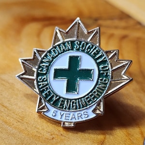 Op de afbeelding: Een gouden en groene pin met een esdoornbladontwerp. De pin heeft een wit kruis met een groene rand en de tekst "Canadian Society of Safety Engineering" en "5 Years".