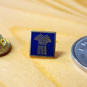 Könnte beinhalten: Eine blaue und goldene Emaille-Anstecknadel mit einem Weizenähren-Design. Die Nadel befindet sich auf einer Holzoberfläche neben einer kanadischen Münze.