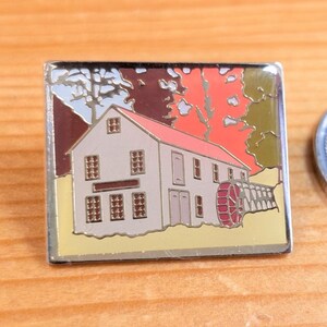 Peut inclure: Une épingle rectangulaire représentant un bâtiment avec un toit rouge et une roue à aubes. Le bâtiment est beige avec des fenêtres et des portes marron. Le fond a des teintes orange, rouge et verte, avec une bordure argentée.