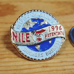May include: A circular enamel pin with a light blue border, featuring the text "Helping Children Is" and "Shriners Business". The pin displays a globe, the word "Nile" in red, the year "1996", and the word "Potentate".