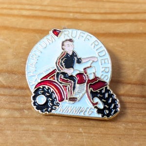Op de afbeelding: Een ronde speld met een persoon op een rode motorfiets. De speld heeft de tekst "KHARTOUM RUFF RIDERS WINNIPEG" in een cirkelvormig ontwerp. De speld heeft een gouden rand en is geplaatst tegen een houten achtergrond.