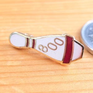 May include: A gold-coloured bowling pin-shaped pin with a white centre and red accents. The pin features the number "800" in gold. The pin is set against a light brown wooden surface.
