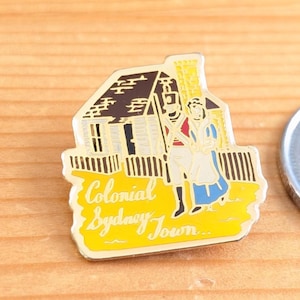 May include: A rectangular enamel pin featuring a colonial scene. The pin depicts a house, two figures in period clothing, and the words "Colonial Sydney Town" in yellow. The pin has a yellow border and is set against a wooden surface.