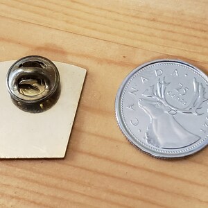 May include: A silver pin with a back showing a pin back and a front showing a design. The pin is next to a Canadian 25 cent coin with a caribou on it.