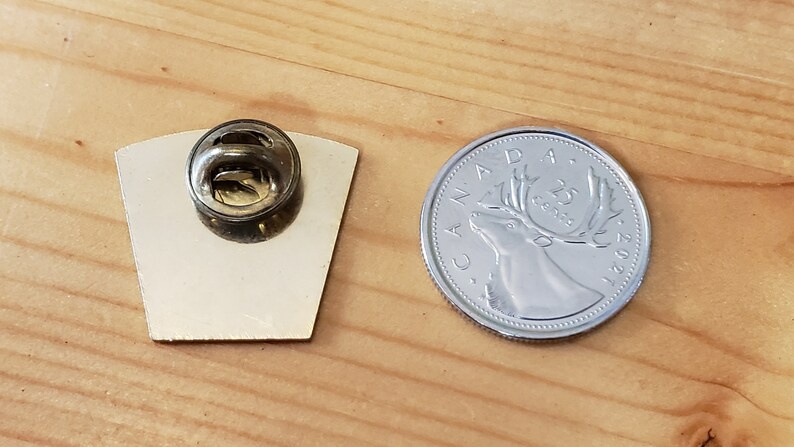 May include: A silver pin with a back showing a pin back and a front showing a design. The pin is next to a Canadian 25 cent coin with a caribou on it.