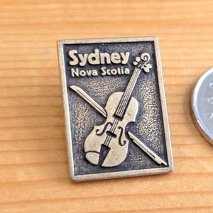 Könnte beinhalten: Eine rechteckige, bronzefarbene Metallnadel mit einem Geigen- und Bogen-Design. Die Nadel trägt die Worte "Sydney Nova Scotia" in einer stilisierten Schriftart. Die Nadel befindet sich auf einer Holzoberfläche.