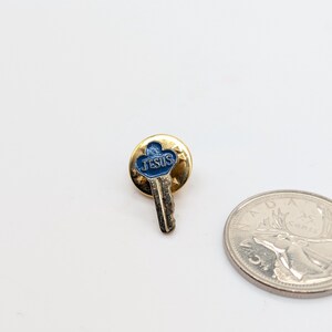 May include: A gold-toned pin featuring a key design with a blue enamel center. The word "JESUS" is inscribed in white. A Canadian 25-cent coin is next to the pin.