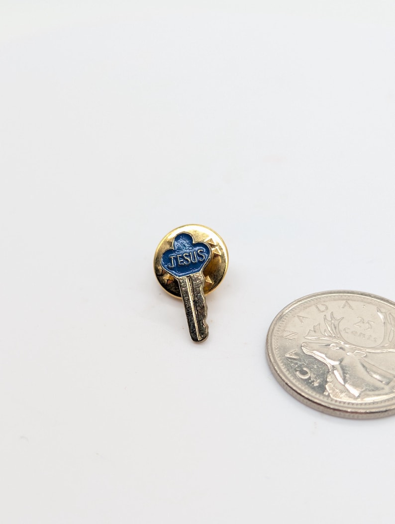 May include: A gold-toned pin featuring a key design with a blue enamel center. The word "JESUS" is inscribed in white. A Canadian 25-cent coin is next to the pin.