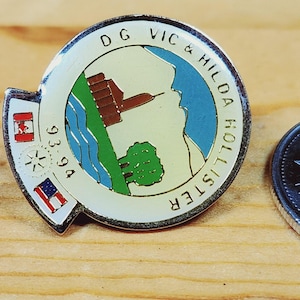 May include: A silver pin with a white background and a colourful image of a building, trees, and water. The text "DG VIC & HILDA HOLLISTER 93 94" is printed around the image.