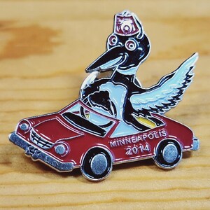 May include: Enamel pin depicting a cartoon bird in a fez driving a red car. The car has the text "MINNEAPOLIS 2014". The bird has black and white feathers and blue wings. A collectible item.