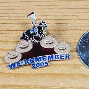 Peut inclure: Épingle émaillée représentant un cavalier sur un cheval noir, un drapeau et quatre chapeaux beiges. L'épingle porte l'inscription "WE REMEMBER 2005" en bleu. Une pièce de monnaie canadienne est visible.