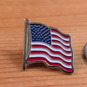 May include: A small, enamelled pin showing the American flag. The pin has a blue field with white stars and red and white stripes. The flag is waving, and the pin has a silver-coloured metal border and pole.