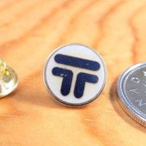 May include: A round, vintage pin with a white background and a navy blue stylized "T" design. The pin has a silver-colored metal border and a pin back. A gold-colored pin and a coin are also visible.