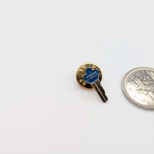 May include: A gold-toned pin in the shape of a key, with a blue enamel top that reads "JESUS". The pin is set against a white background, with a quarter coin visible.