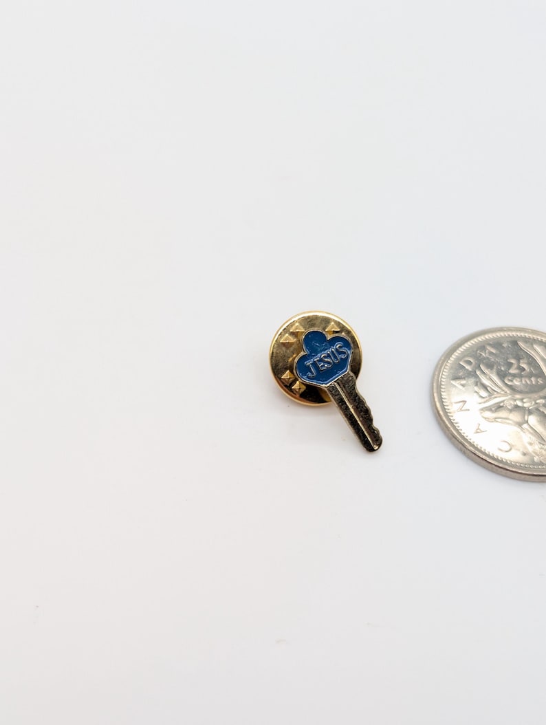 May include: A gold-toned pin in the shape of a key, with a blue enamel top that reads "JESUS". The pin is set against a white background, with a quarter coin visible.
