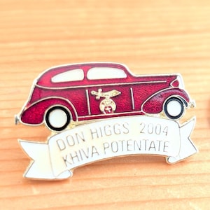 May include: A burgundy enamel car-shaped pin with white detailing. The pin reads "DON HIGGS 2004 KHIVA POTENTATE" on a white banner. The car has a gold emblem. A vintage-style collectible.