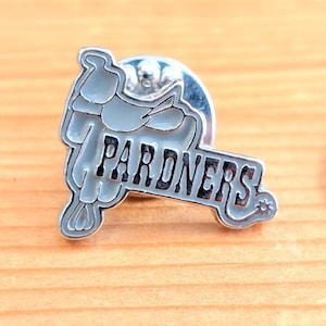 May include: A silver-coloured pin featuring a light grey saddle design and the word "PARDNERS" in black lettering. The pin has a star detail and a silver pin back.