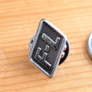 Könnte beinhalten: Ein silberfarbener Ansteckpin mit den Buchstaben "F" und "7" in einem rautenförmigen Rahmen. Der Pin hat einen geriffelten Hintergrund und ein klassisches Design, geeignet für Jacken, Taschen oder Hüte.
