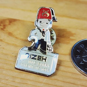 May include: A vintage metal pin featuring a cartoon character in a red fez and white coat, standing on a rectangular base. The base has the text "IZEH" and "L.R. MAC BARRY POTENTATE 1987". A coin is next to the pin.