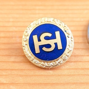 Op de afbeelding: Een ronde, goudkleurige speld met een blauw midden. Het midden heeft een goudkleurig monogram. De speld heeft een decoratieve, sierlijke rand. De speld ligt op een lichtbruin houten oppervlak.