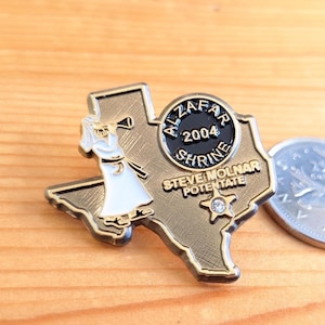 Puede incluir: Un pin dorado con la forma del estado de Texas. El pin presenta un emblema circular con el texto "AL ZAFAR SHRINE 2004" y "STEVE MOLNAR POTENTATE". Una figura con túnica blanca toca una trompeta. También hay una pequeña gema en forma de estrella.