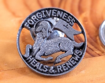 May include: A silver-toned round pin with the words "FORGIVENESS HEALS & RENEWS" encircling a lamb carrying a banner with a cross. The pin is set against a wooden surface.