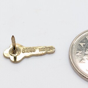 May include: A gold-colored key-shaped pin with the text "CROSS KOREA" is displayed. The pin has a pointed post for attachment. A silver coin is partially visible in the image, providing a size reference.