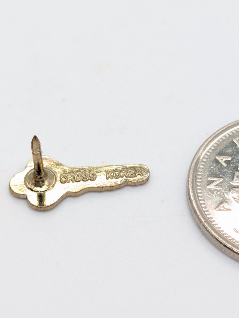 May include: A gold-colored key-shaped pin with the text "CROSS KOREA" is displayed. The pin has a pointed post for attachment. A silver coin is partially visible in the image, providing a size reference.