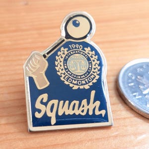 Op de afbeelding: Een vintage Squash-speld met een gouden rand en een marineblauwe achtergrond. De speld toont een squashracket, het woord "Squash" in goud en de tekst "1990 International Games Edmonton".