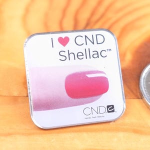 May include: A square, silver-edged product package with the text "I ❤️ CND Shellac™" and a pink nail polish design. The CND logo and the words "Hands. Feet. Beauty." are also visible.