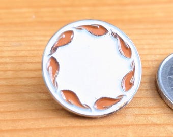May include: A round, white metal pin with a silver border. The pin features a ring of orange leaf-shaped designs around the edge. The pin is set on a light brown wooden surface.