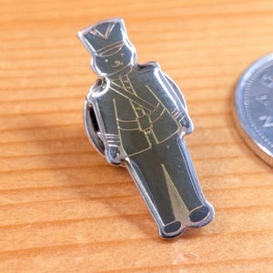 May include: A silver-toned metal pin in the shape of a toy soldier. The pin features a dark green uniform with gold accents and a black hat. The pin is on a wooden surface.